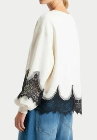 Maglione bianco con bordo in pizzo nero a scallop all'orlo e alle maniche. Texture morbida, vestibilità ampia, abbinato a denim azzurro chiaro.