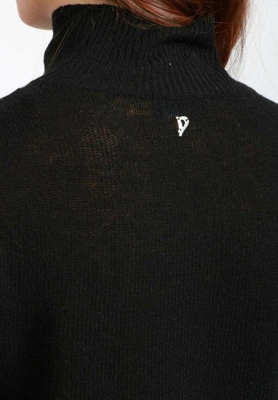 Donna con i capelli rossi che indossa un maglione a collo alto nero a costine, caratterizzato da un piccolo dettaglio in argento a forma di lettera "D" sulla parte superiore della schiena.