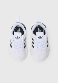 Άσπρα αθλητικά παπούτσια με υφή στην μύτη, μαύρες τρεις ρίγες, κα�ι αντίθετο μαύρο ταμπάκι στη φτέρνα. Σχεδιασμός με κορδόνια και λογότυπο adidas.