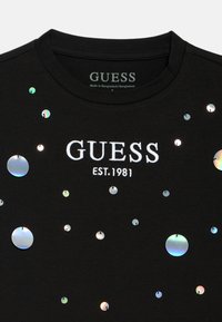 Μαύρο κοντομάνικο μπλουζάκι με τη φράση "GUESS EST. 1981" κεντημένη με λευκό νήμα, διακοσμημένο με ιριδίζουσες στρογγυλές παγιέτες διαφόρων μεγεθών.
