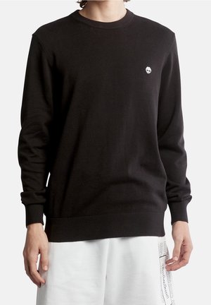 Person trägt einen schwarzen Timberland Langarmpullover mit kleinem weißem Logo auf der Brust und weiße Shorts mit aufgedrucktem Text.