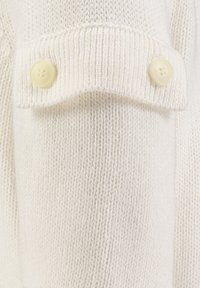 Witte gebreide trui met textuurstof. Voorzien van een opgerolde mouw met twee lichtgekleurde knopen. Evenwichtige, gladde steken overal.