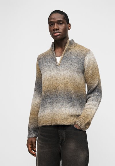 Άνδρας που φοράει πολύχρωμο γκρι, μπεζ και μπλε πλεκτό πουλόβερ με κοντό γιακά με φερμουάρ και σκουρόχρωμο ξεβάμμενο τζιν.
