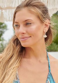 Silver shell-shaped earrings hang from the model's ear. She has long, wavy blonde hair and is wearing a multi-colored halter bikini top.