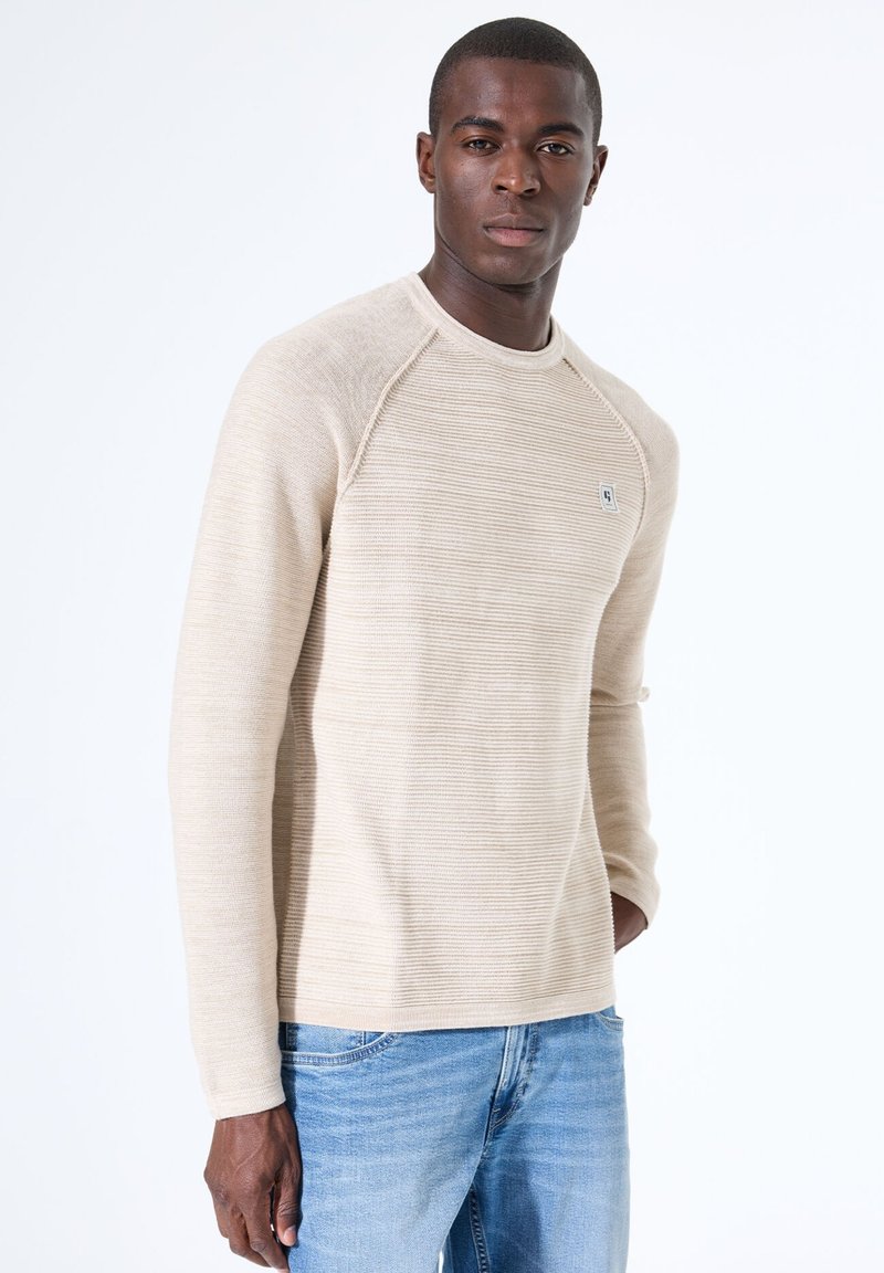 Beige geribde lange mouwen trui met een ronde halslijn en raglanmouwen, gecombineerd met lichtblauwe jeans. Subtiele branding op de borst.