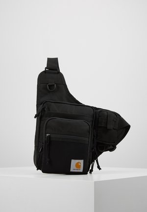 Sac bandoulière noir Carhartt avec plusieurs poches zippées et une sangle ajustable, présenté sur un piédestal blanc sur fond gris.