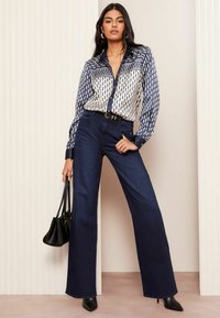 Bluse mit Marine-Muster und geometrischem Design, kombiniert mit weit geschnittenen dunklen Denim-Hosen. Die Accessoires umfassen eine schwarze Handtasche und spitze Absätze.