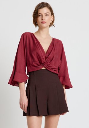 Donna che indossa una camicetta rossa scuro con nodo intrecciato e maniche larghe, abbinata a pantaloncini plissettati marrone scuro a vita alta.