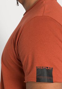 Camiseta de manga corta color óxido hecha de algodón. Presenta un logo negro "REPLAY" en la manga izquierda y un cuello acanalado. Textura suave.