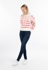 Musterpullover mit pastellrosa Herzformen auf einem cremefarbenen Hintergrund, kombiniert mit dunkelblauen Jeans und weißen Turnschuhen.