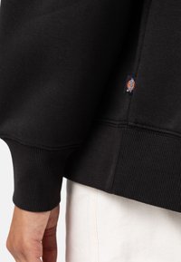 Black sweatshirt with a ribbed cuff and hem, featuring a small branded tag on the side. White textured fabric is partially visible.