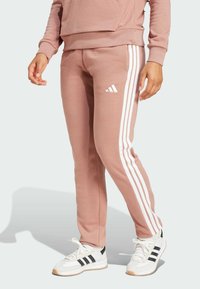 Růžové tepláky s bílými proužky Adidas po stranách, vyrobené z měkké látky. Nosit s bílými teniskami s černými akcenty.