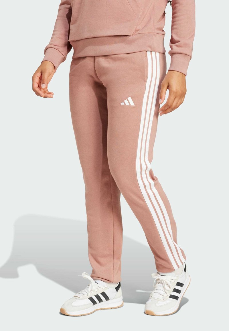Růžové tepláky s bílými proužky Adidas po stranách, vyrobené z měkké látky. Nosit s bílými teniskami s černými akcenty.