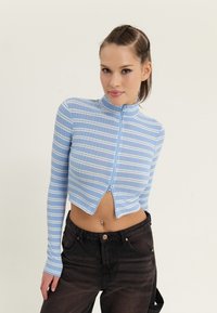 Lyseblå glidelås-kort topp med hvite striper, lange ermer og høy krage; paret med svarte høytaljede jeans.
