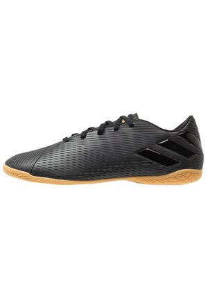 Chaussure de football en salle noire avec tige texturée, lacets noirs, trois bandes brillantes sur le talon et semelle en caoutchouc marron clair.