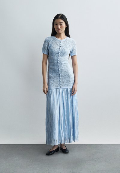 Νεαρή γυναίκα που στέκεται, φορώντας ένα ανοιχτό μπλε δαντελωτό φόρεμα με κουμπιά μπροστά και πλισέ φούστα, συνδυασμένο με μαύρα φλατ παπούτσια, μπροστά σε απλό φόντο.