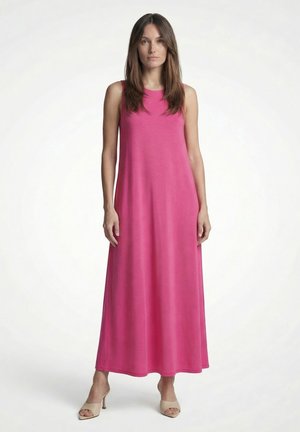 Frau steht, trägt ärmelloses, bodenlanges rosa Kleid und beige offene Absätze vor weißem Hintergrund.