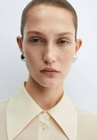 Camicia beige con colletto e chiusura a bottoni, indossata con grandi orecchini rotondi. La pelle è chiara e i capelli sono pettinati all'indietro. Illuminazione morbida.