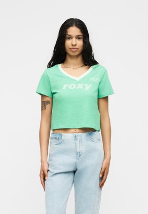 Jeune femme aux longs cheveux foncés, portant un t-shirt vert court à col en V avec le texte "roxy" et un jean taille haute bleu clair, debout immobile.