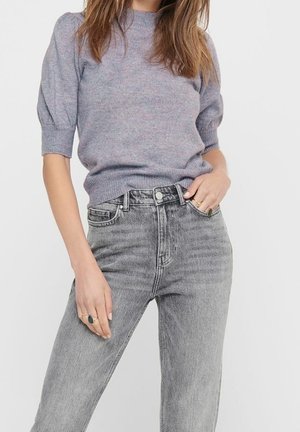 Femme portant un haut en maille à manches courtes violet clair rentré dans un jean gris délavé taille haute, avec des bagues aux deux mains.