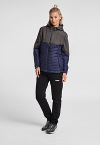 Vadderad jacka med grå övre del och marinblå nedre del, med blixtlåsstängning och huva. Kombinerad med svarta byxor och grå skor med texturerade sulor.