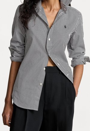 Femme portant une chemise à rayures verticales noires et blanches avec manches retroussées et pantalon noir ample, une main dans la poche.