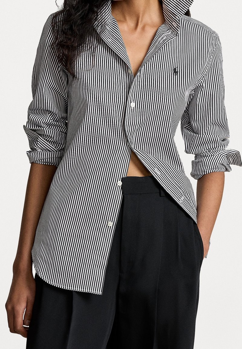 Femme portant une chemise à rayures verticales noires et blanches avec manches retroussées et pantalon noir ample, une main dans la poche.