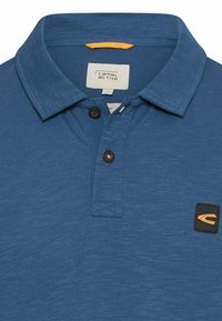 Blauw poloshirt met twee zwarte knopen, camel active-label aan de binnenkant van de kraag, en een klein zwart patch met geel gebogen logo op de borst.