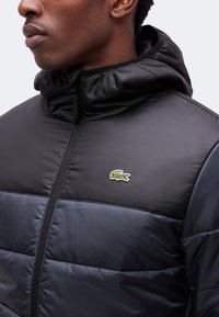 Homme portant une doudoune noire et bleu marine avec capuche, fermeture éclair fermée, avec un petit logo crocodile vert sur la poitrine.