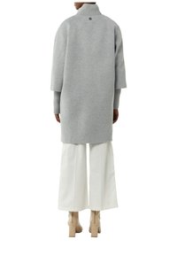 Grau wollartiger Mantel mit hohem Kragen, gerippten Ärmelbündchen und einem Knopfdetail auf der Rückseite. Kombiniert mit weit geschnittenen weißen Hosen und beigen Ankle Boots.