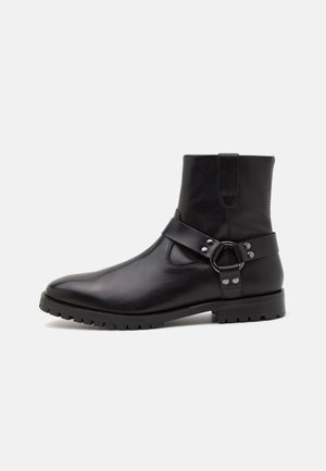 Czarne skórzane botki za kostkę z szpiczastym noskiem, płaską gumową podeszwą oraz dekoracyjnym paskiem z metalowym pierścieniem i ćwiekami. Gładka faktura.