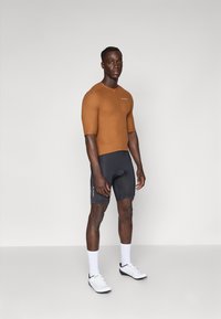 Brun tilpasset sykkeltrøye med korte ermer, frontglidelås, kombinert med svarte sykkelshorts, hvite sokker og hvite sportssko.