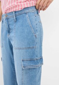 Lichtblauwe denim cargobroek met zijzakken, voorzien van zichtbare stikseldetails en een relaxte pasvorm, weergegeven vanaf de zijkant met een gestreept top.