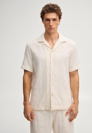 Jonge man met een gestructureerd off-white kort mouwen overhemd met knopen en bijpassende korte broek tegen een effen achtergrond.