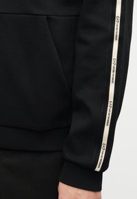 Μαύρο μανίκι με λευκή ταινία EA7 Emporio Armani κατά μήκος του βραχίονα και το χέρι εμφανές στο κάτω άκρο.