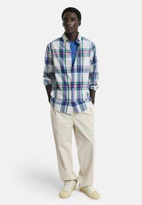 Camisa de cuadros abotonada en blanco, azul, verde y púrpura. Combinada con pantalones de pierna ancha de color crema y zapatillas beige. Textura suave.