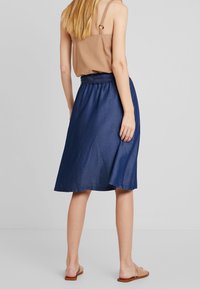 Nainen, joka on pukeutunut vaaleanruskeaan hihattomaan puseroon, sinisiin polvipitkiin hameeseen ja beigeihin sandaaleihin, seisoo tasaisella harmaalla taustalla selkäpuolelta.