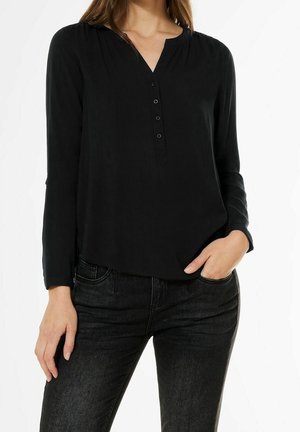 Vrouw draagt een zwarte, lange mouw, met knopen, blouse ingestopt in zwarte denim jeans, met één hand in de zak en de andere ontspannen langs haar lichaam.