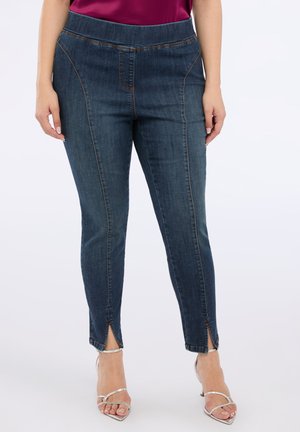Dark blue denim pants with a high waistband, featuring stitched detailing, side pockets, and ankle slits. Paired with silver strappy heels.