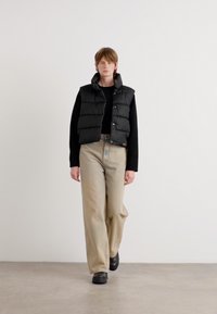 Colete acolchoado preto com colarinho alto, usado sobre uma blusa de manga longa, combinado com calças largas bege e sapatos pretos. Fundo liso.
