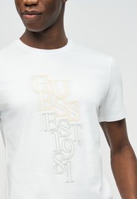 Άνδρας που φορά λευκό T-shirt με κατακόρυφο, επικαλυπτόμενο κείμενο "GUESS EST 1981" τυπωμένο μπροστά σε ανοιχτό μπεζ και γκρι γράμματα.