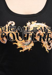 Μαύρο τοπ με στρογγυλή λαιμόκοψη, διακοσμημένο με το κείμενο "VERSACE JEANS COUTURE" πάνω από περίτεχνους χρυσούς και λευκούς μπαρόκ στροβιλισμούς με παγιέτες.