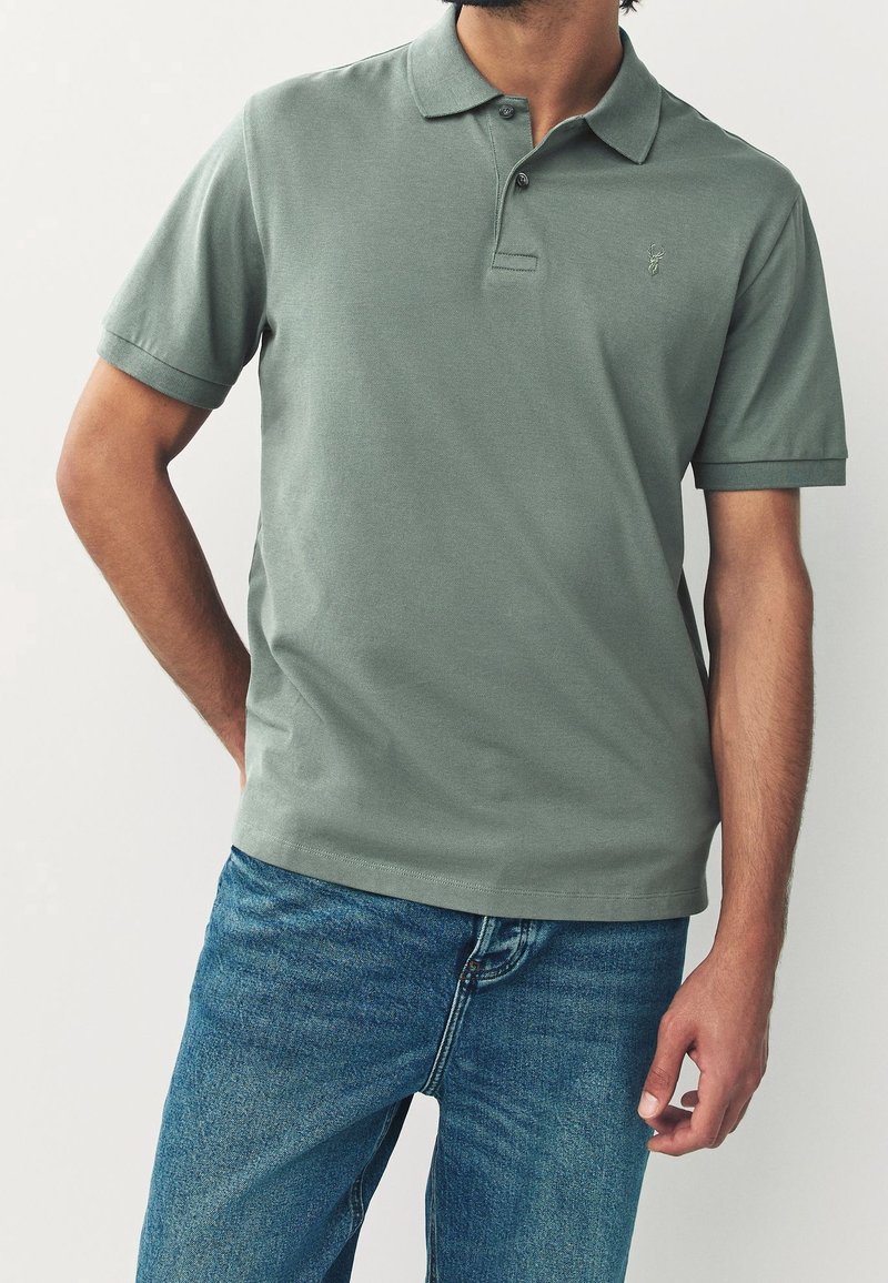 Homme portant un polo vert atténué à manches courtes et un jean bleu, debout avec une main derrière le dos.