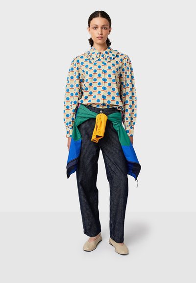 Musterhemd in Beige mit blauen und roten Punkten, grün und gelb geschichtete Jacke an der Taille gebunden, gestreifte marineblaue Hose und beigefarbene Schuhe.