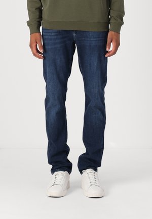 Donkerblauwe denim jeans met een rechte pasvorm, gecombineerd met witte sneakers en een olijfgroen sweatshirt. Gladde textuur over de gehele stof.