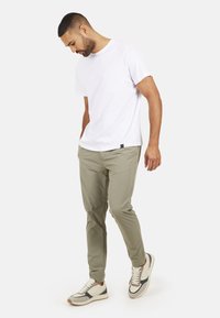 Biała bawełniana koszulka, jasne zielone spodnie i beżowe trampki z szarymi akcentami. Model pozuje w luźnej pozycji, z rękoma w kieszeniach, patrząc w dół.