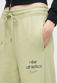 Άτομο που φοράει ανοιχτοπράσινο παντελόνι φόρμας Nike Athletics με ελαστική μέση και κορδόνι, με το χέρι στην πλαϊνή τσέπη, συνδυασμένο με ένα μαύρο κοντό τοπ.