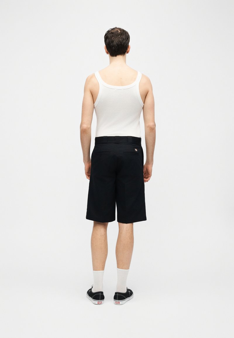 Uomo con canottiera bianca, pantaloncini neri fino al ginocchio, calzini bianchi e scarpe da ginnastica nere, in piedi di schiena su sfondo bianco uniforme.