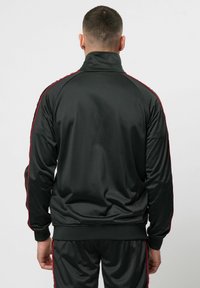 Zwarte trainingsjack met hoge kraag, rode zijkanten, gladde stof en ribgebreide manchetten. Geen zichtbare branding of aanvullende accenten.