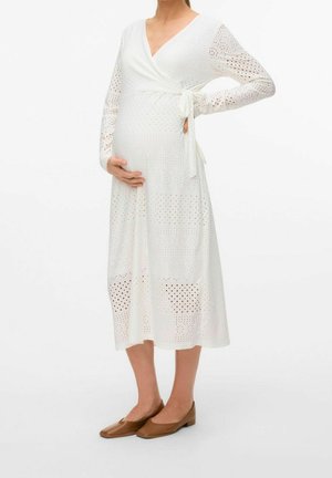 Personne enceinte portant une robe portefeuille midi blanche perforée et des ballerines marron, tenant son ventre avec une main sur la hanche.